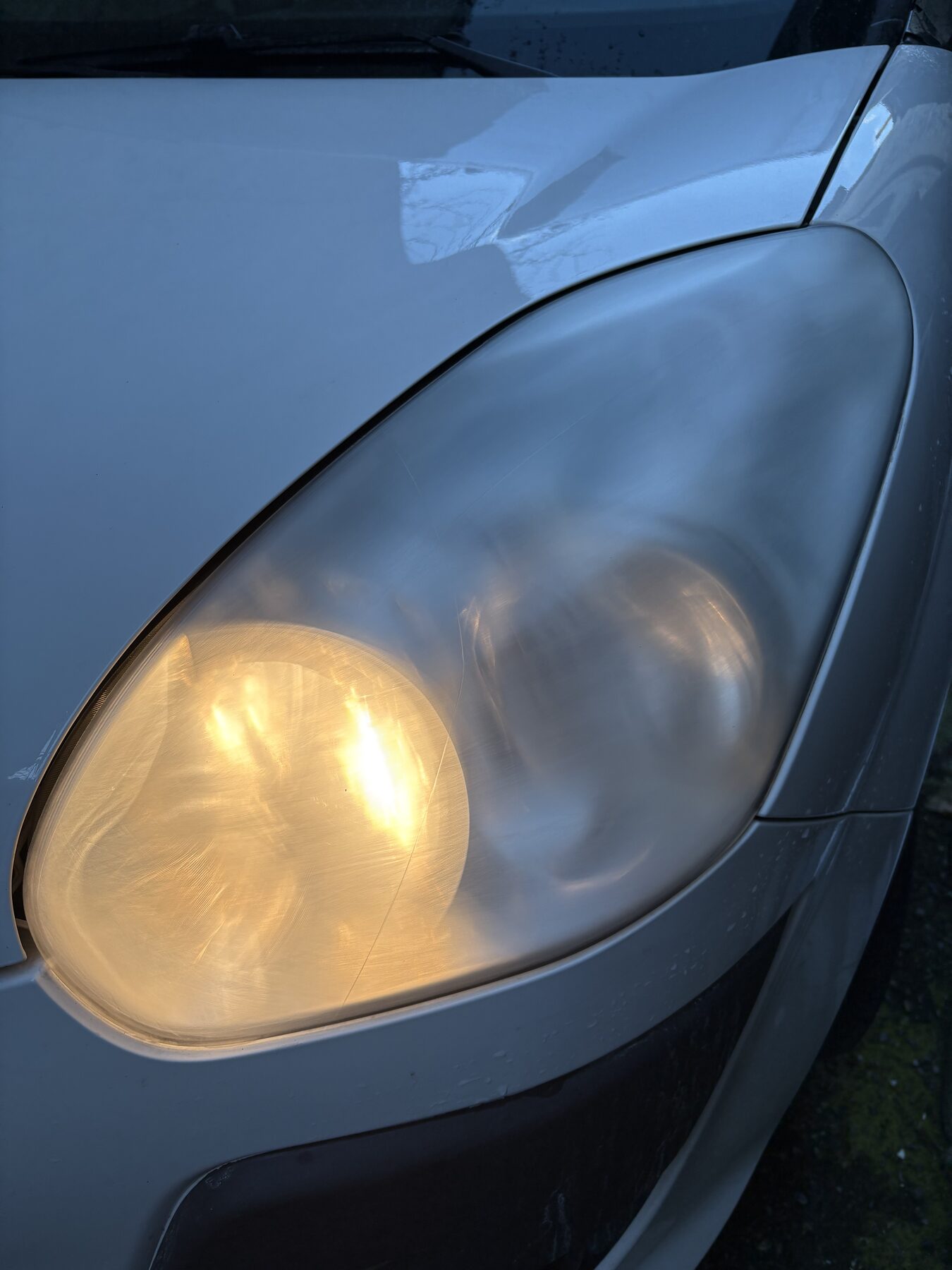 Faro anteriore opaco di un'utilitaria grigia, con la luce accesa